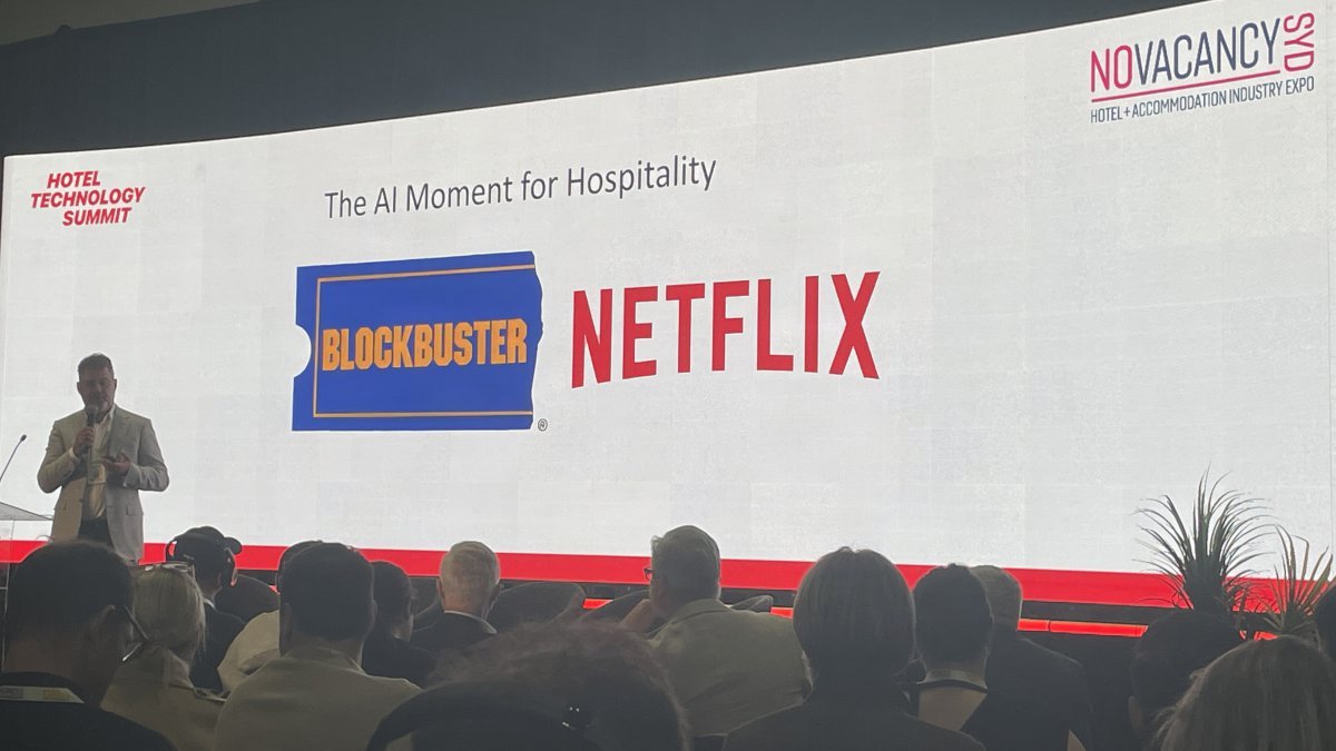 Presenter beside a slide titled The AI Moment for Hospitality at NoVacancy Sydney 2026, featuring Blockbuster and Netflix graphics.